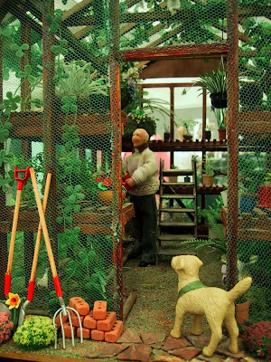 One-twelfth scale miniature green house with a man in a hand-knitted jumper working inside and a dog watching from the doorway.