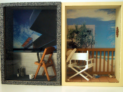 Two one-twelfth scale modern miniature balcony scenes in box frames. On the left is a city balcony, with sprayed concrete walls and floor and a corrugated iron balcony wall. On the balcony is a folding wooden chair with a laptop on it. On the floor is a cordless phone, a mug and a stovetop espresso jug. On the beach balcony is a white director's chair with a woven hat on the back, a magazine on the seat, and a pair of jandals and a glass on the floor. The balcony overlooks the sea and some cabbage trees.